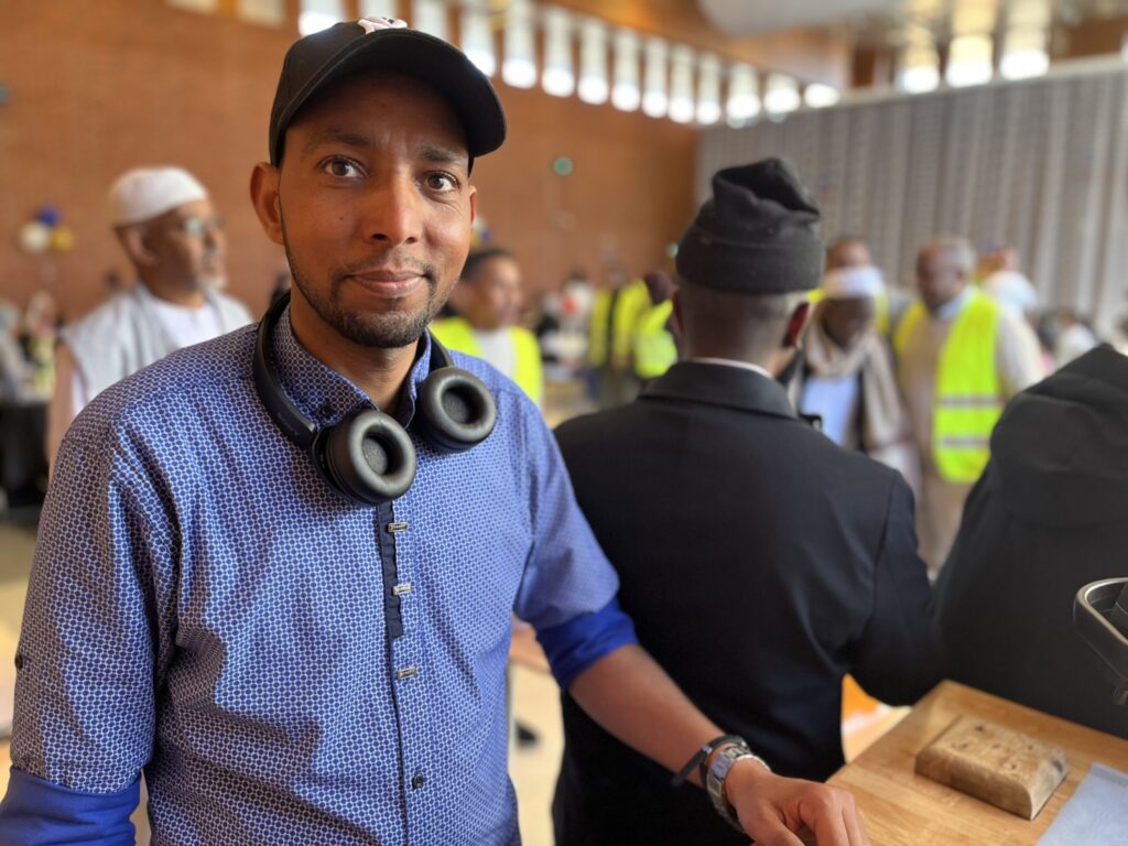 man med keps och blå skjorta ler mot kameran framför festligheter 