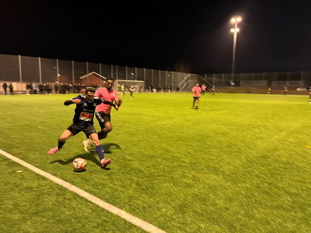 Fotbollsspelare i svart matchtröja dribblar bollen från en i rosa matchtröja.