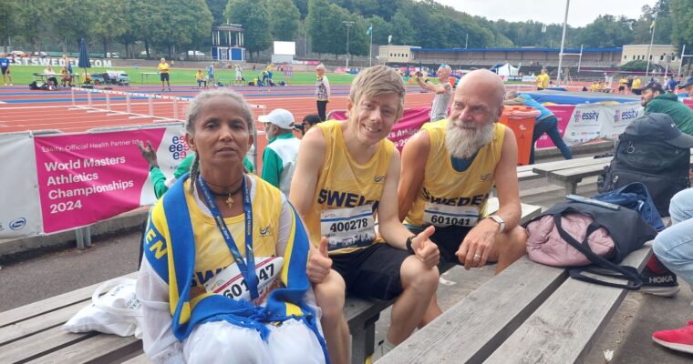 en kvinna och två män med sportkläder för Sverige sitter på en bänk framför en friidrottsarena