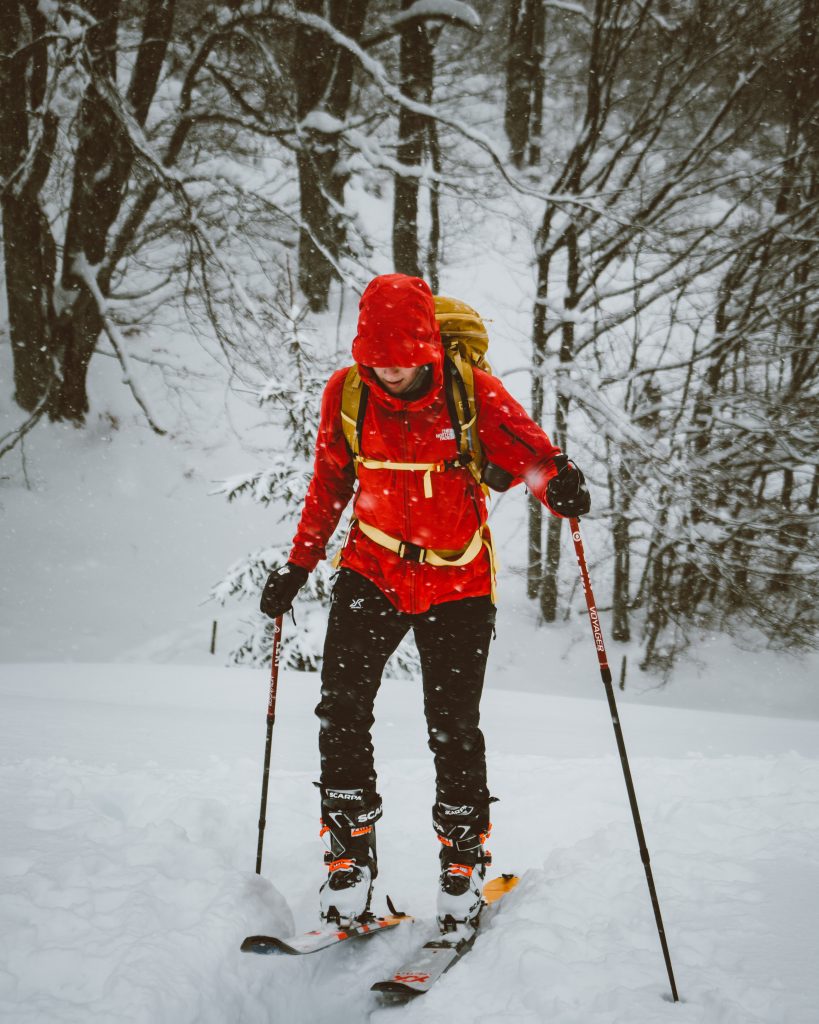 Skitour, Skitourengehen, Frau mit Skiern, in schwarzer Hose mit einer roten Jacke, verschneite Berge im Hintergrund. Die Sonne scheint, perfektes Wetter, winterliche Stimmung.