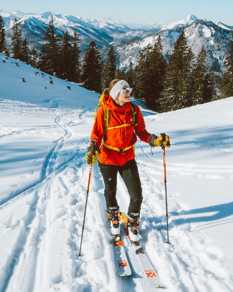 Skitour, Skitourengehen, Frau mit Skiern, in schwarzer Hose mit einer roten Jacke, verschneite Berge im Hintergrund. Die Sonne scheint, perfektes Wetter, winterliche Stimmung.