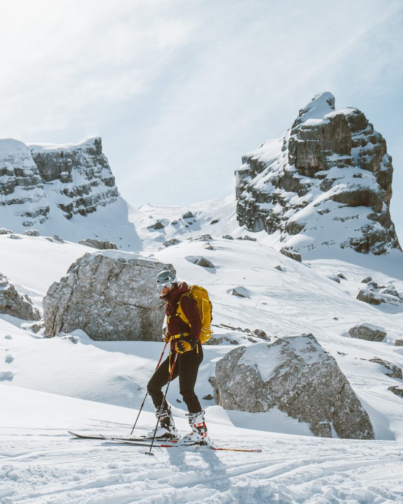 Skitour, Skitourengehen, Frau mit Skiern, in schwarzer Hose mit einer dunkelroten Jacke, verschneite Berge im Hintergrund. Die Sonne scheint, perfektes Wetter, winterliche Stimmung.