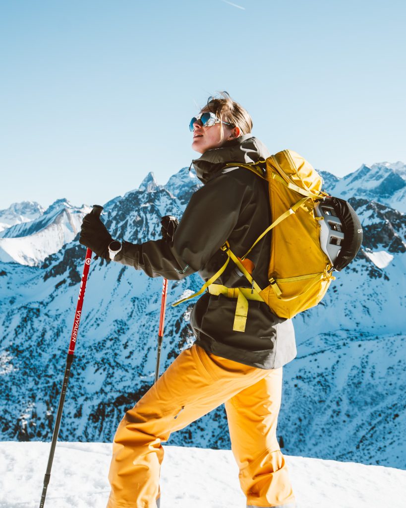 Skitour, Skitourengehen, Frau mit Skiern, in orangener Hose mit einer grünen Jacke, verschneite Berge im Hintergrund. Die Sonne scheint, perfektes Wetter, winterliche Stimmung.