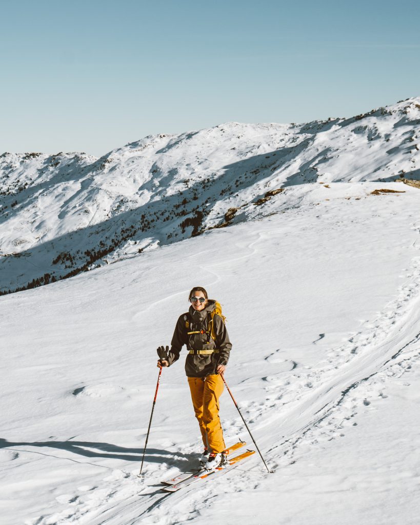 Skitour, Skitourengehen, Frau mit Skiern, in orangenen Hose mit einer grünen Jacke, verschneite Berge im Hintergrund. Die Sonne scheint, perfektes Wetter, winterliche Stimmung.