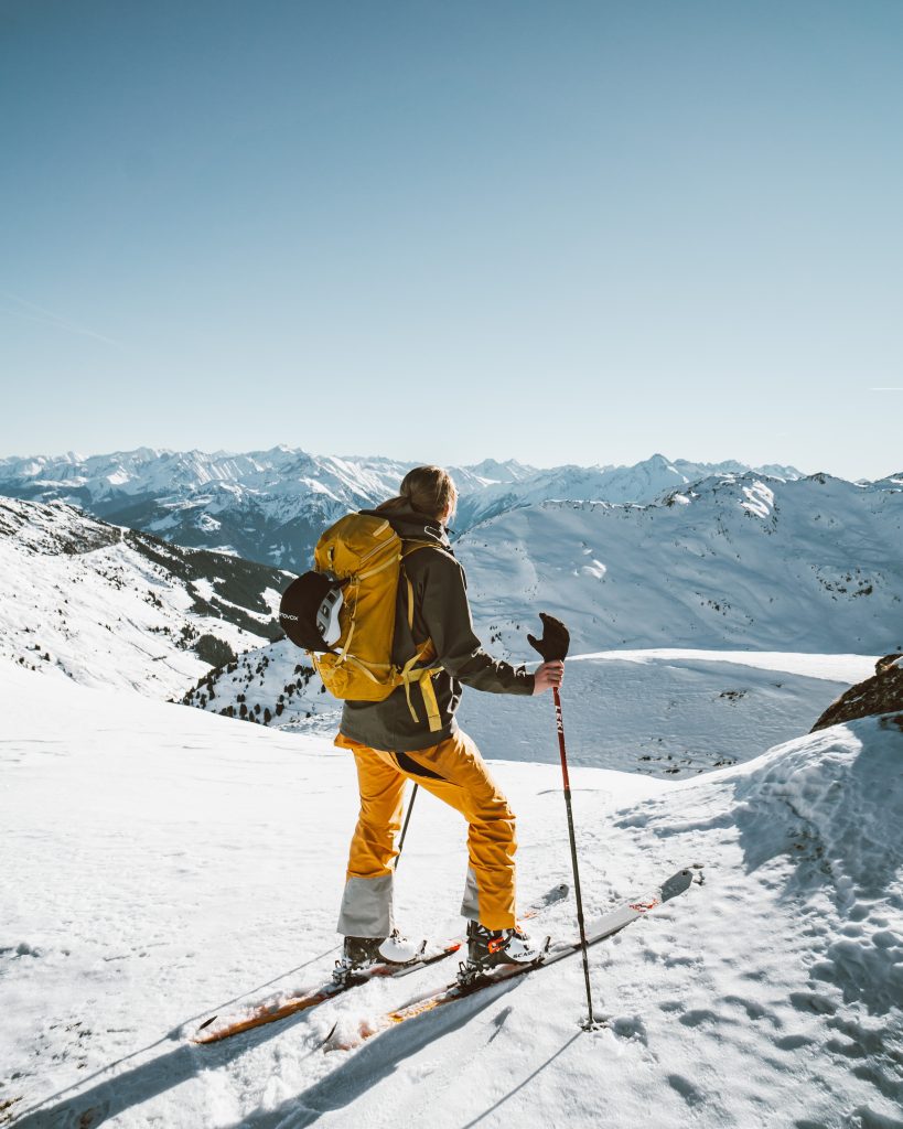 Skitour, Skitourengehen, Frau mit Skiern, in orangener Hose mit einer grünen Jacke, verschneite Berge im Hintergrund. Die Sonne scheint, perfektes Wetter, winterliche Stimmung.