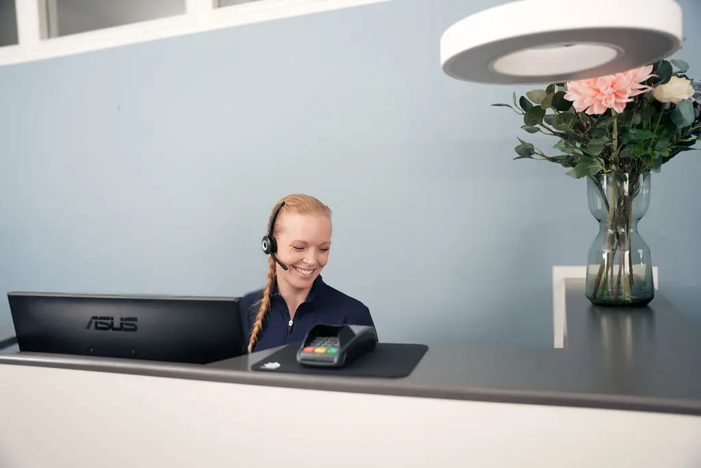 En smilende receptionist taler i headset bag skranken og tager imod henvendelser.