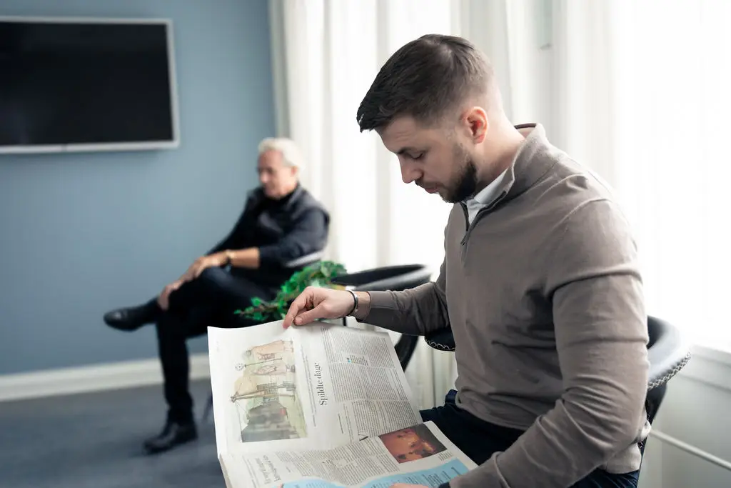 To patienter slapper af i venteområdet. Den ene læser avis, mens den anden hviler sig.