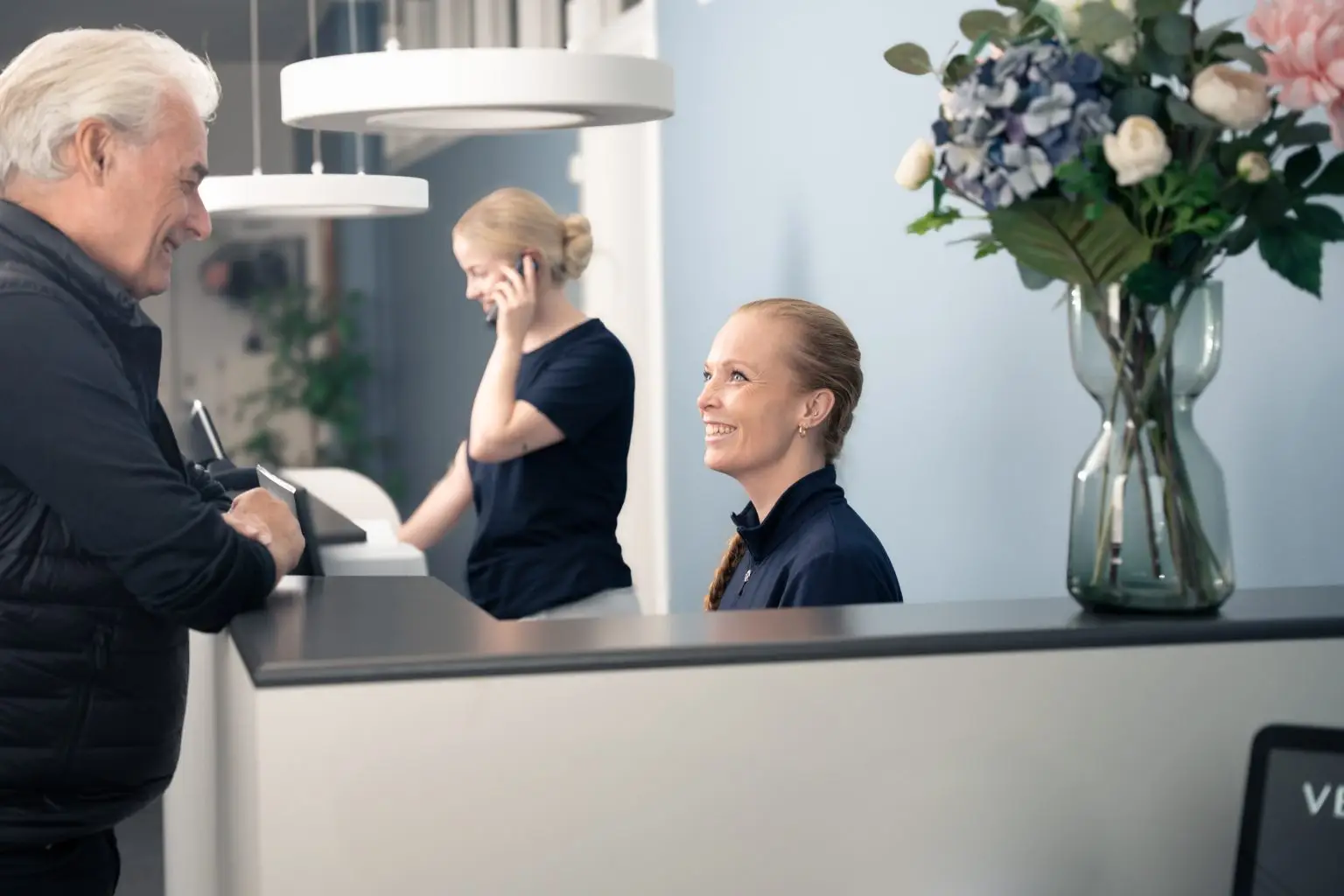 Patient bliver venligt modtaget af smilende personale i receptionen hos tandlægerne Ølandshus på Amager. En anden medarbejder hjælper en patient over telefonen.