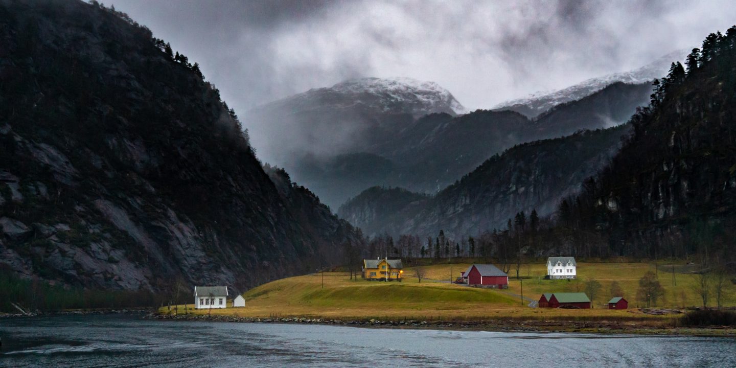 Bilde av en gård med tilhørende bygg nær vannet på Vestlandet. Fjell i bakgrunnen.