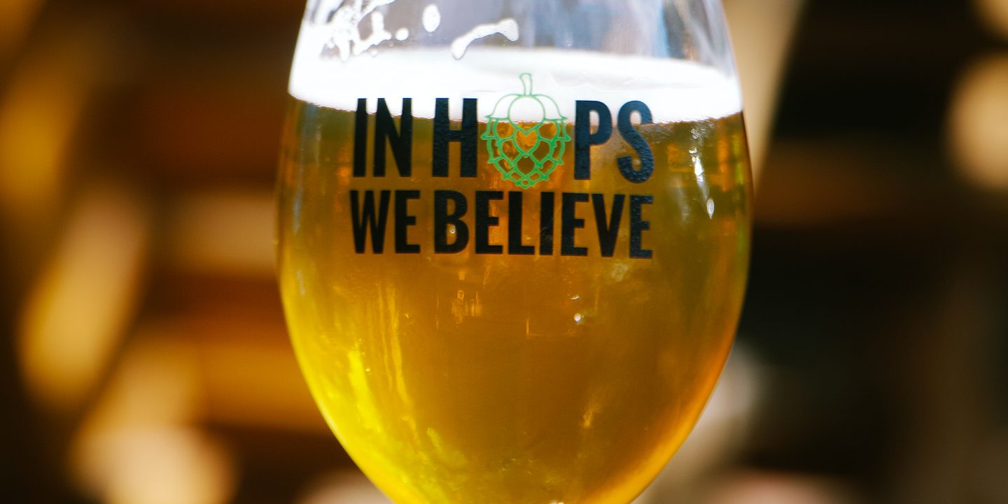 Et glass på bordet fylt med øl. Glasset har teksten "In Hops We Believe"