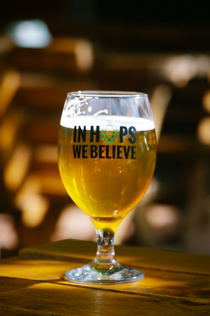 Et glass på bordet fylt med øl. Glasset har teksten "In Hops We Believe"