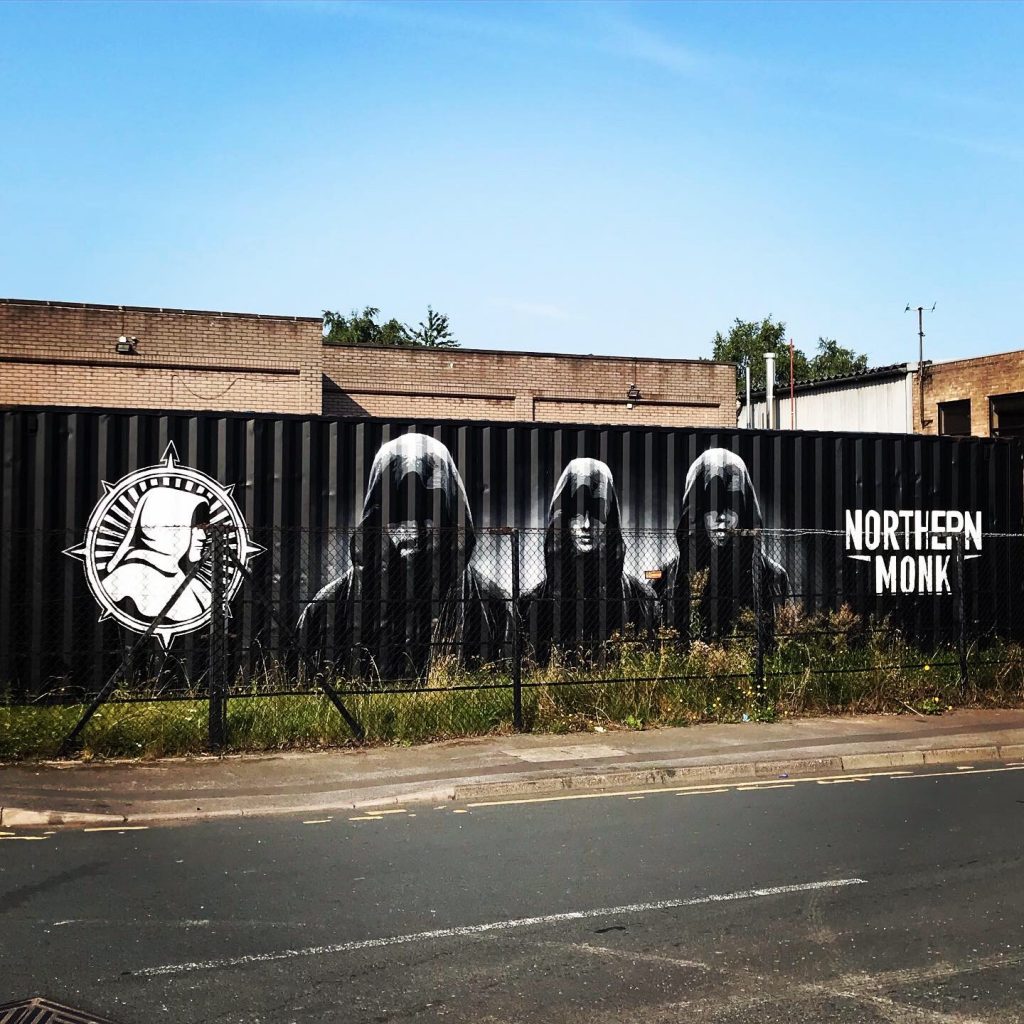 En svart container i Leeds med logoen til Northern Monk og bilde av tre skumle munker. Blå himmel. Foran bryggeriet.