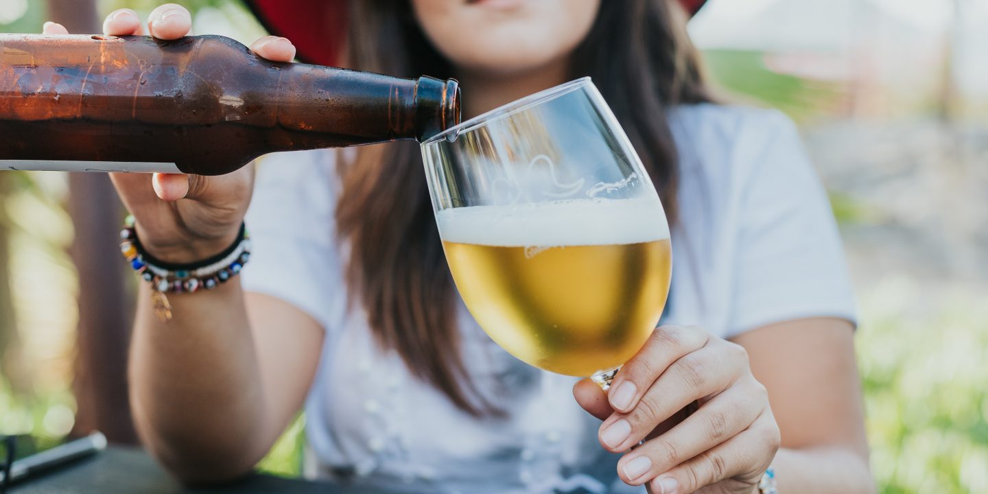En kvinne med hatt som fyller øl fra en brun flaske i et glass med stett.