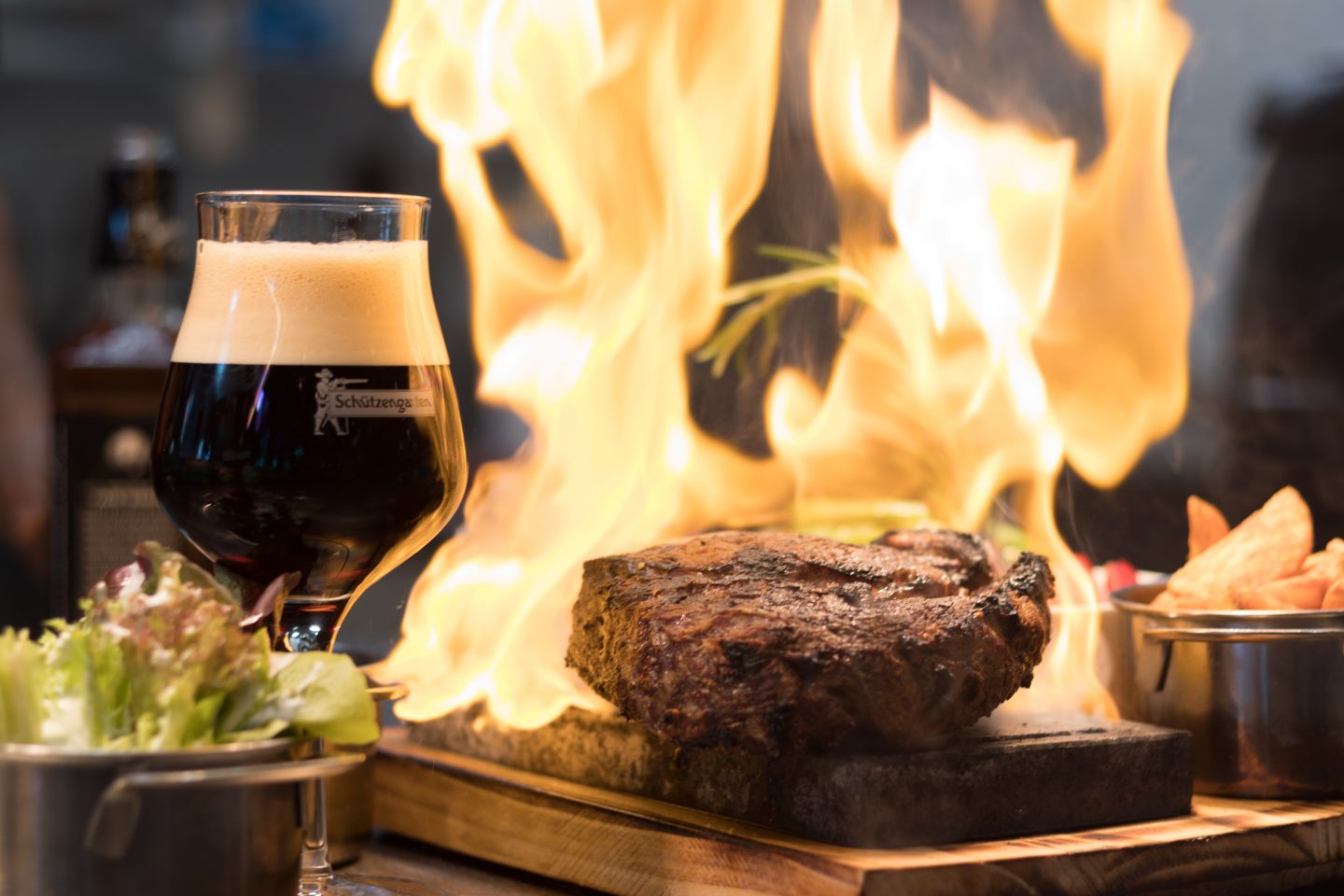 Et glass med mørkt øl i til venstre på bordet. En biff i midten og flamme bak.