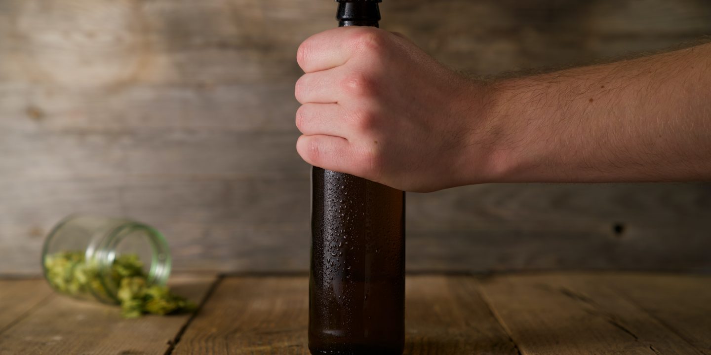 En fyr holder en flaske med øl. Et glass med humleblomster i bakgrunnen.