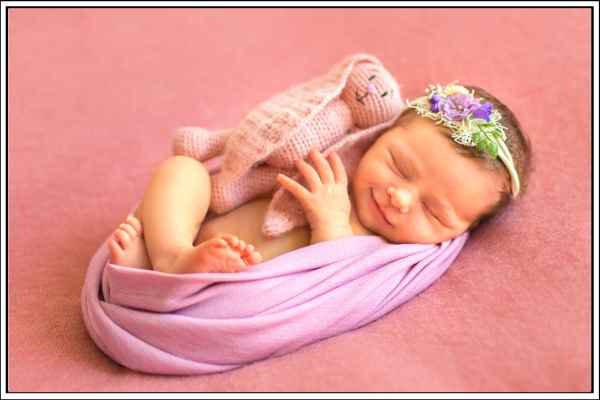 swaddled newborn baby with a smile on her face, and a pink bunny teddy right next to her.