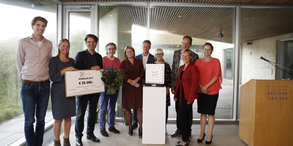 De winnaars van de prijs met organisatie van de prijs samen op een foto met de check en de bloemen