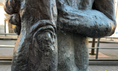 Een detail van een foto van een beeld waarbij je een rug ziet waar iemand zijn arm achterlangs houdt en met zijn hand de elleboog van zijn andere arm vasthoudt