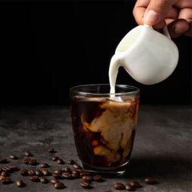Mælk fra kande hældes i et klart glas med kaffe