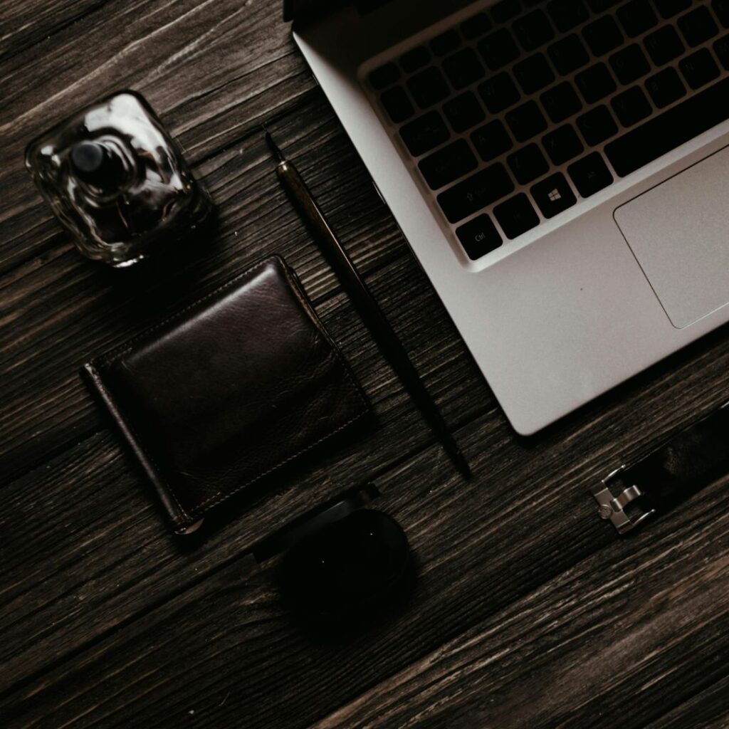 Mysk parfym, elegant plånbok, Macbook dator, dyr penna, snygg klocka och trådlösa hörlurar på ett mörkt träbord.