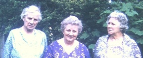 Three elderly women standing together in a leafy garden, wearing patterned blouses and smiling at the camera.