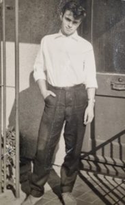 Person standing outdoors, hands in pockets, wearing a white blouse and dark jeans beside a metal railing; black-and-white photo.