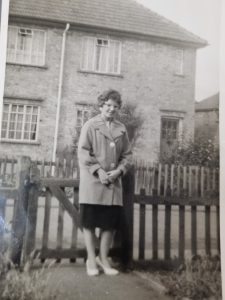 Woman in a light coat standing beside a wooden picket fence in a brick suburban yard, black-and-white photo.