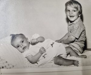 Two young children on a bed in a vintage black-and-white photo; one lies on its side in a light outfit, the other sits up smiling.