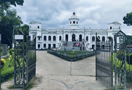 White neoclassical palace with a central dome, seen through open wrought-iron gates on a gravel path; people gather on the steps.