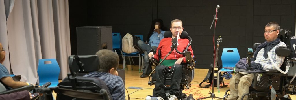 CEO, Jon Rey-Hastie presents to a room. He is in an electric wheelchair and wearing a red jumper