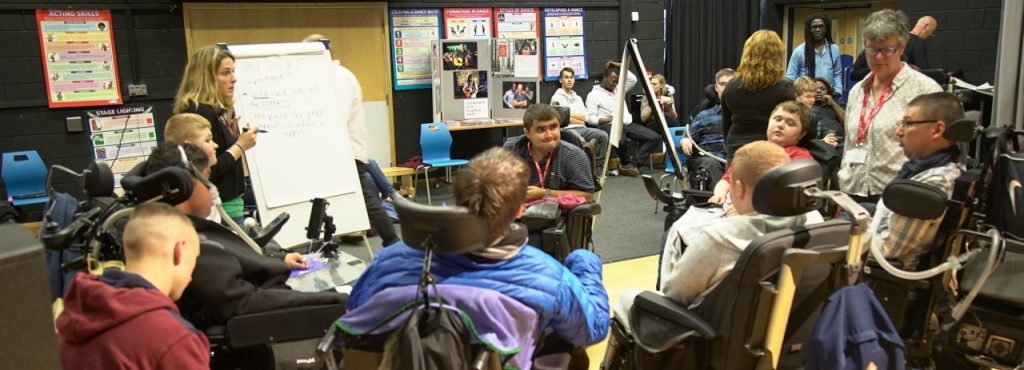 A group of people at a Pathfinders event. There are people in powerchairs engaging in conversation and a female standing at a flipchart.