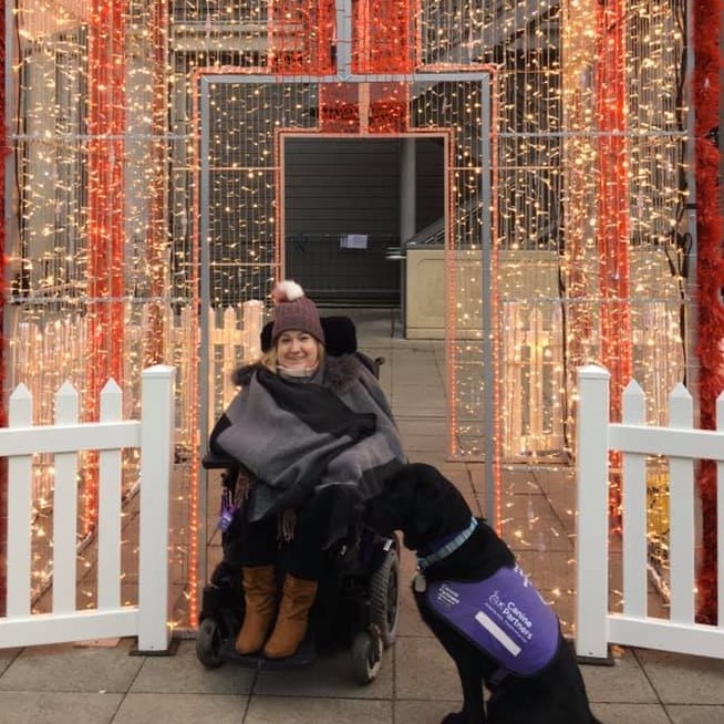White woman with blonde hair and brown eyes in a wheelchair, wearing a grey coat and brown bobble hat posing with a large black dog in front of white fairy lights.