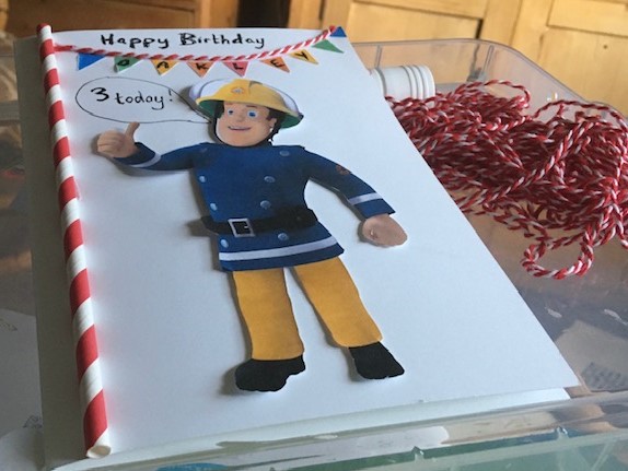 Handmade card with cut out fireman sam in the centre. Yellow helmet, blue jacket and yellow trousers. A red and white candy striped straw along the left side.