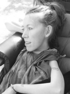 Black and white photo of the head and shoulders of a white woman sat in a chair looking to the left. Her hair in a high ponytail