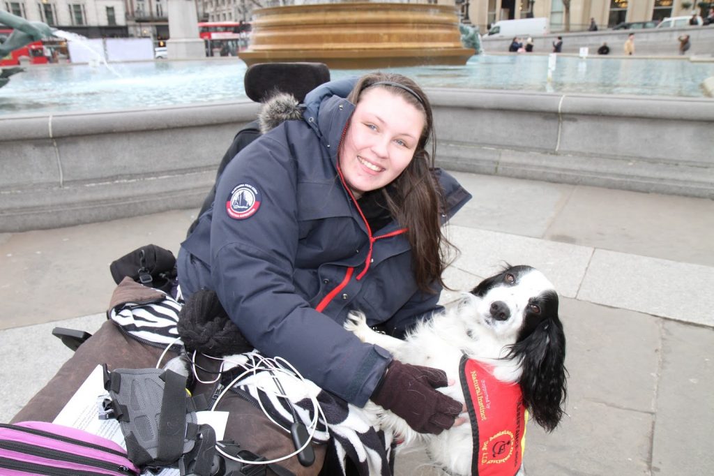 White female wheelchair user with brown hair, brown eyes, smiling, wearing navy coat, cuddling small white and black dog in front of grey fountain.