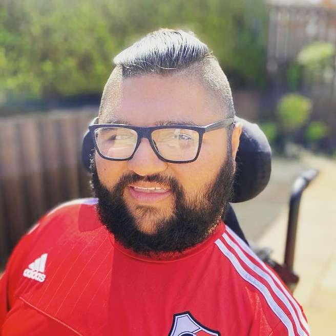 Head and shoulders of a young man with full beard and mustache with short dark hair and black rimmed glasses. Sat in a wheelchair in the garden wearing a red football shirt