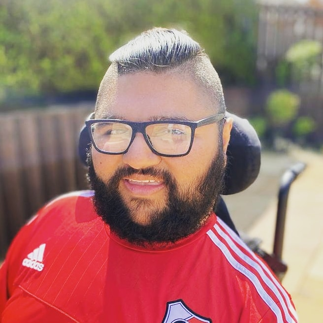 Head and shoulders of Sanjeev Mann, a young man with full beard and mustache with short dark hair and black rimmed glasses. Sat in a wheelchair in the garden wearing a red football shirt.