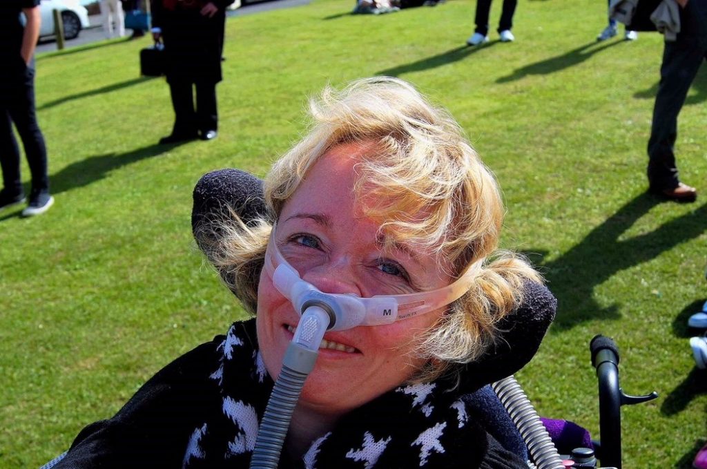 Picture of Sarah Rose, a blonde woman smiling on some grass, a ventilator mask over her nose. Sarah is a trustee and co-chair of trustees at Pathfinders