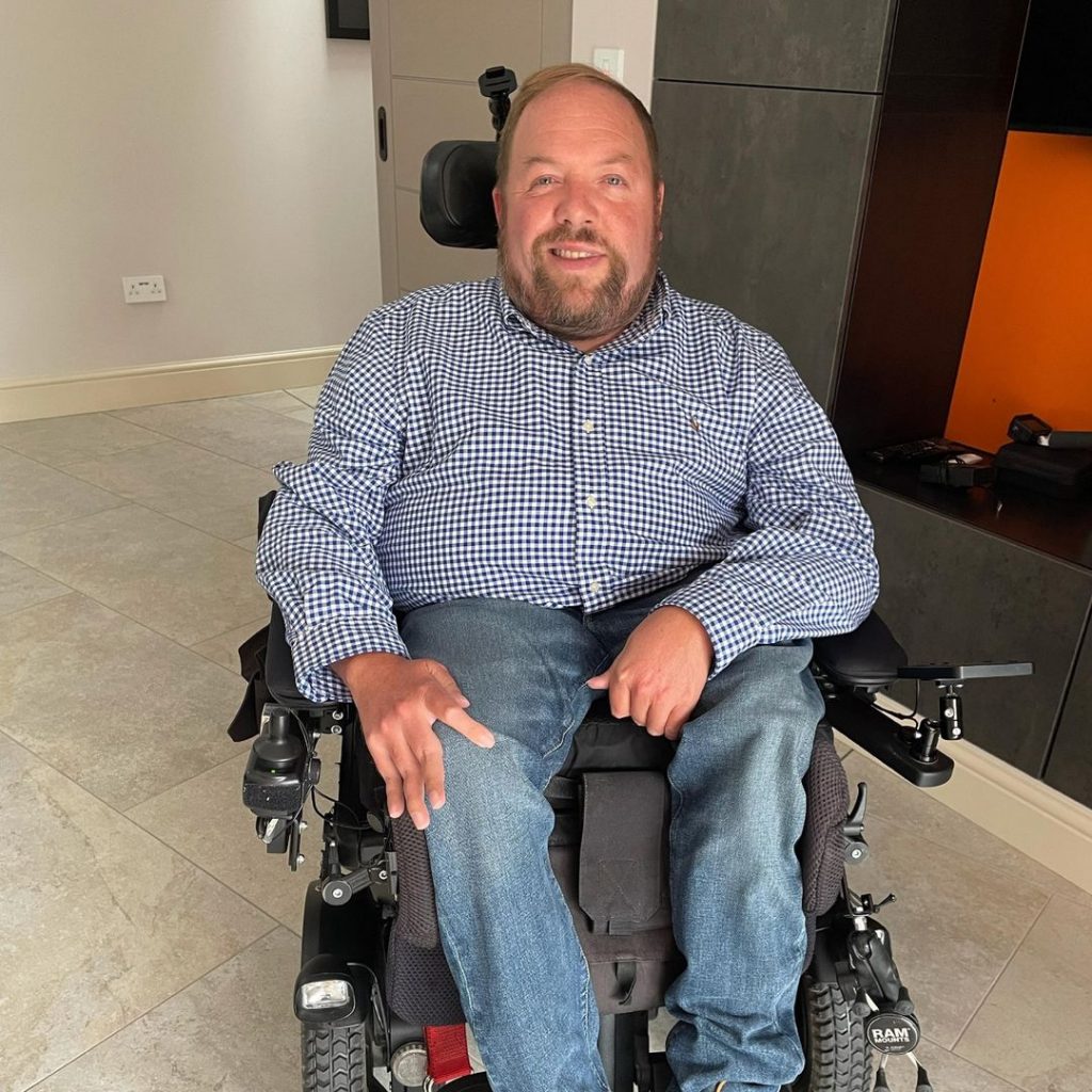 A man with brown hair sitting in a powered wheelchair in a house