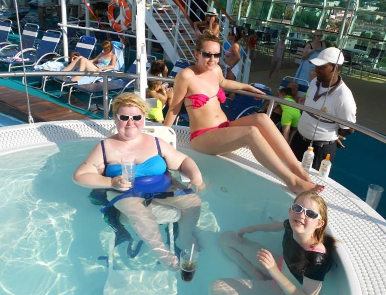 Blond haired woman in a blue swimsuit wearing sunglasses sat on pool hoist in a hot tub on a cruise ship deck holding a drink. A woman in a pink bikini sat on the side of the hot tub and young girl in the hot tub wearing a black t shirt and sun glasses.