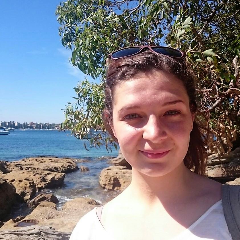 A picture of Olivia Morgan, a white woman, against a background of trees, blue sky, and water