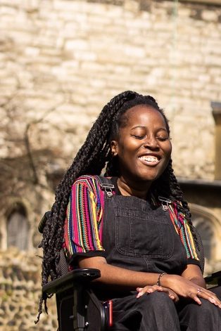 Jacqui, a black woman sits in an electric wheelchair in front of an old castle, laughing with her eyes closed.