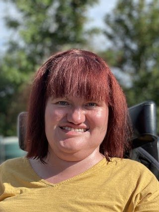 Michaela Hollywood, a white woman in an electric wheelchair, wearing a mustard top with red hair