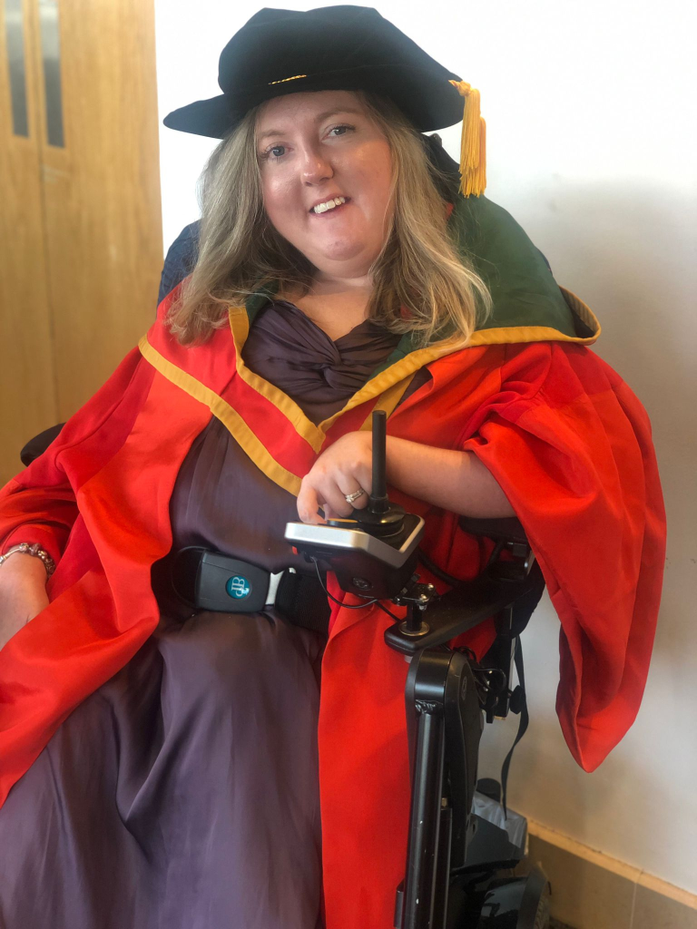 Suzanne Glover, a white woman wearing red graduation robes, in an electric wheelchair