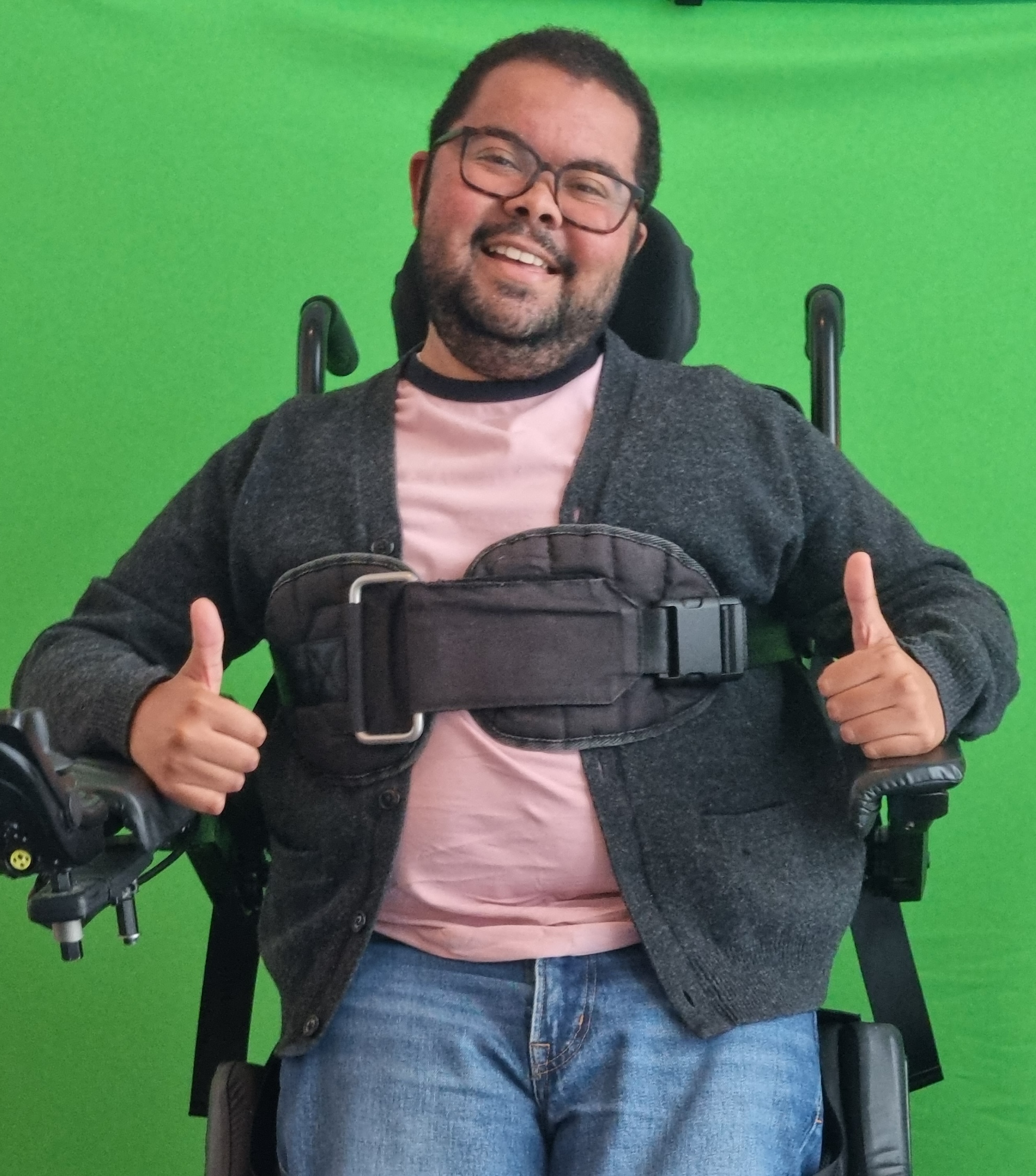 A photograph of Benjamin James in a standing powerchair in front of a green screen giving two thumbs up.
