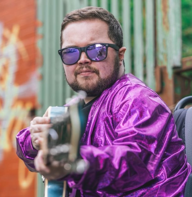 Alex, a white male with short brown hair. He is wearing a shiny purple jacket and sunglasses while holding a guitar.