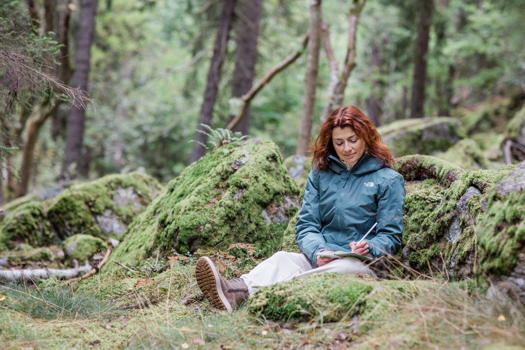 a calm photo of a woman writing in a notebook while sitting in a mossy forest in Stockholm