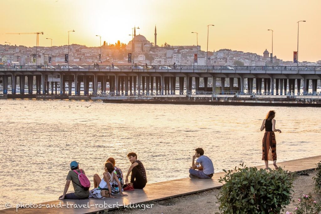 Young turkish people are sitting at the shore of Golden Horn in front of Atatürk Bridge in Istanbul
