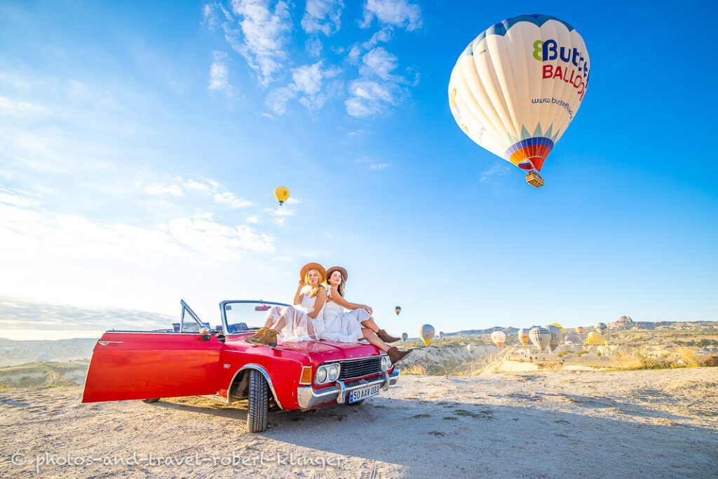 Two instagram travelling girls sitting on a red ford car in Cappadocia with hot air ballons in the background