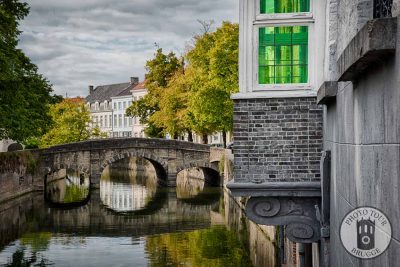 A green Venetian-styled glass panel sits by an arched bridge on a canal in Bruge Belgium. Photo by Photo Tour Brugge.